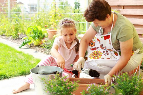 A woman and a young girl are working together outdoors in a well-maintained garden, engaging in gardening activities; the woman is wearing a floral apron and gloves, while the girl is dressed in casual clothing with pink and white stripes. They are tending to potted plants placed on a paved patio area bordered by a wooden fence. The garden features a neatly trimmed lawn with lush green grass, vibrant flower beds with blooming plants, and a variety of shrubs and small trees in the background. The scene captures a sunny day, with natural light illuminating the garden and casting gentle shadows, indicative of pleasant weather. The setting appears to be a private residential outdoor space, suitable for gardening and outdoor maintenance services offered by Hedge Trimming Gunnersbury, located in the Gunnersbury area in West London, with a focus on lawn care, hedge trimming, and landscape upkeep.