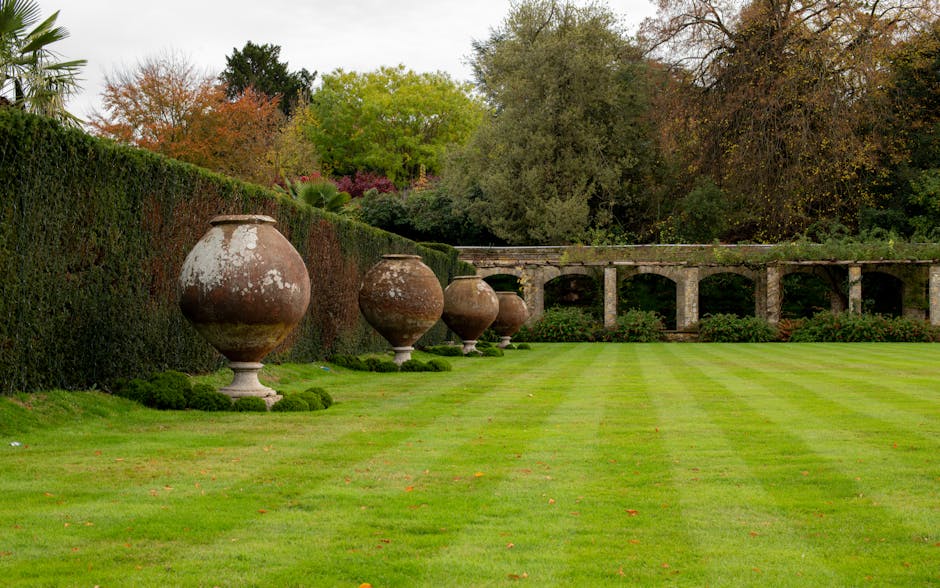 A well-maintained garden featuring a lush, freshly mown lawn with alternating light and dark green stripes. Along the left side, a neatly trimmed hedge runs parallel to a row of large, weathered terra